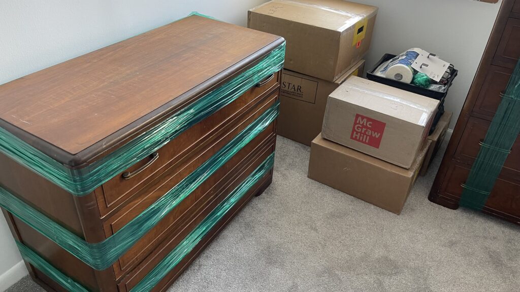 Packed dresser wrapped in moving plastic with cardboard boxes stacked in a bedroom during a local move.