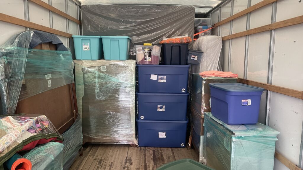 Large furniture wrapped in moving blankets and plastic alongside storage bins in a moving truck.